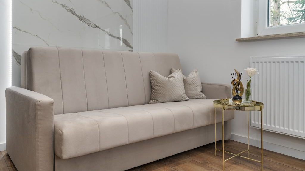 Beige sofa bed with vertical channel tufting beside a small gold side table in a bright modern room.