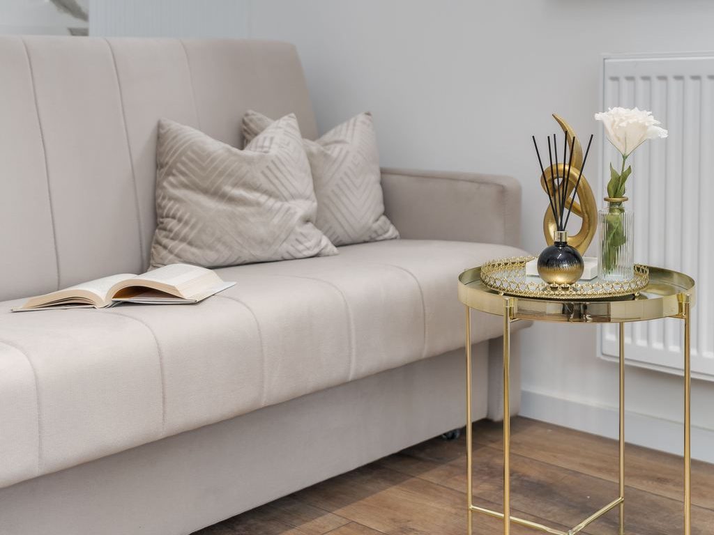 Neutral living room with a beige sofa, gold side table, wood flooring, white walls, and simple decorative accents.