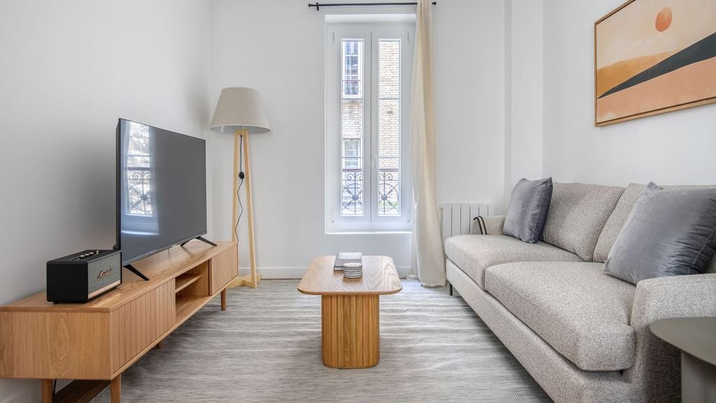 Small modern living room with a gray sleeper sofa, wood TV stand, narrow coffee table, and floor lamp by tall windows.