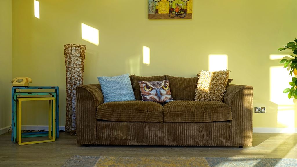 Dark olive-brown corduroy sofa in a sunlit living room with colorful accent pillows, nesting side tables, and a green wall.