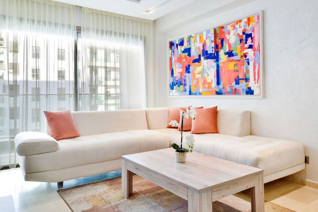 Bright modern living room with a white sectional sofa, coral accent pillows, colorful abstract wall art, and sheer curtains.