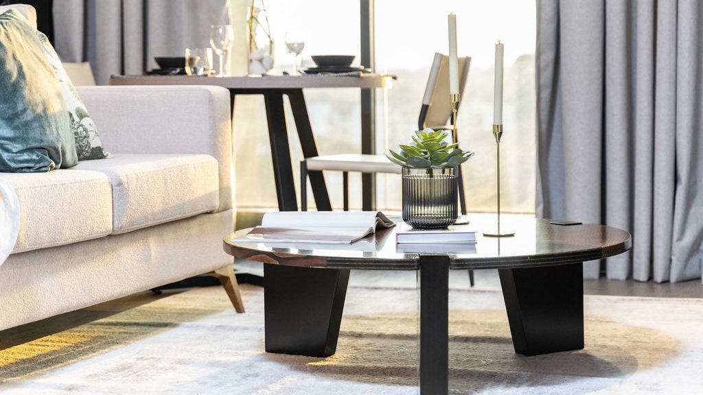 Black round coffee table styled with a small plant, books, and tall candleholders beside a light sofa in a sunlit living room.