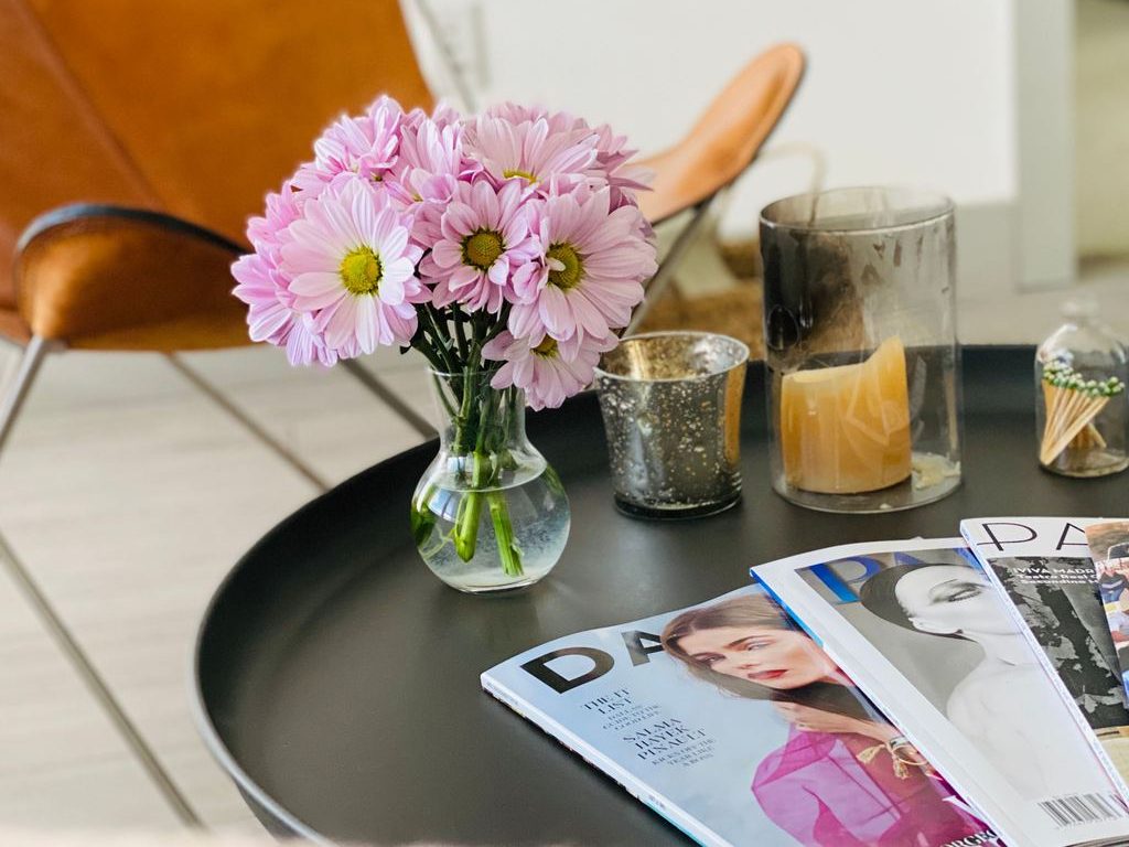 Black round coffee table styled with pink flowers, candles, and fashion magazines beside a leather lounge chair.