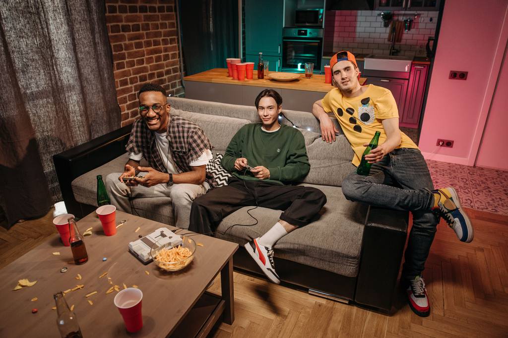 Three friends playing console video games together on a sofa with snacks and drinks nearby