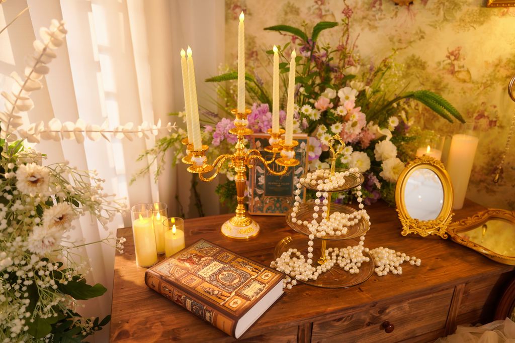 Vintage candle decor on a wooden dresser with gold candelabra, flowers, pearls, books, and warm ambient lighting.