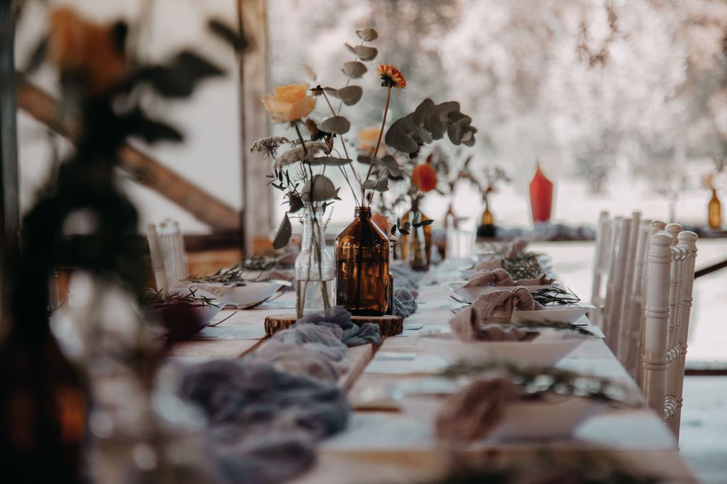 Rustic dining table set with amber glass bottles, loose wildflowers, and layered place settings by a sunlit window.