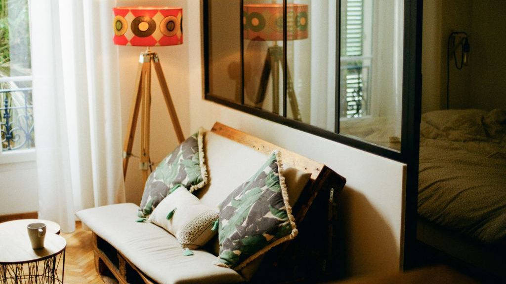 Compact sofa bed styled with patterned pillows beside a tripod floor lamp in a small multipurpose room.