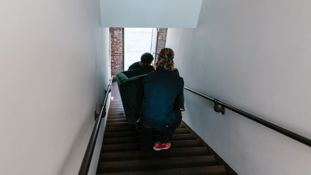 Two people carrying a green sofa down a narrow apartment staircase.