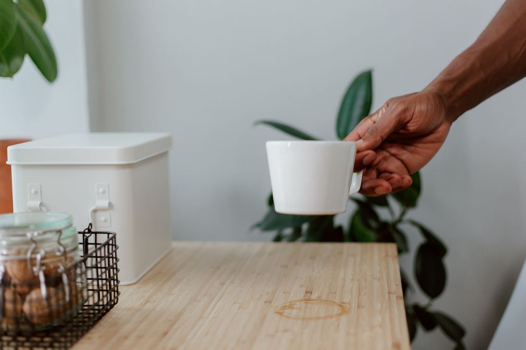 Hand lifting a white mug from a light wood table with a visible circular water ring.