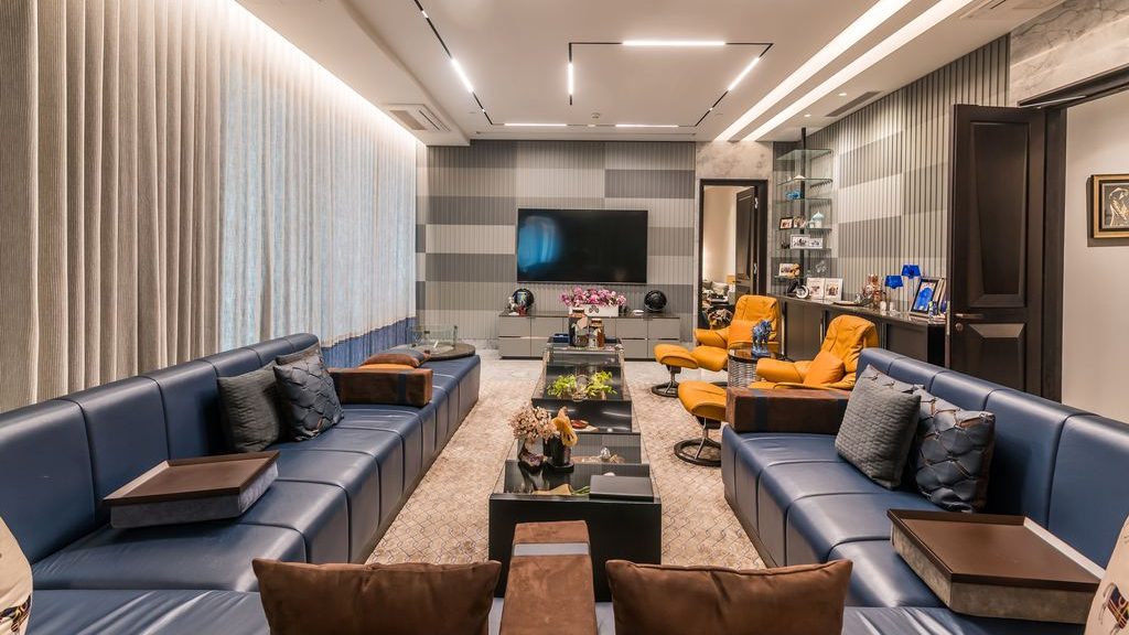 Two blue leather sofas and orange lounge chairs arranged around central coffee tables in a modern conversational living room with a TV.