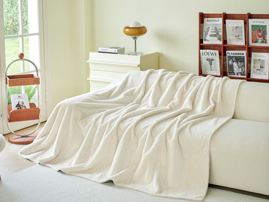 Cream sofa bed draped with a soft white throw beside magazine racks and a small lamp in a bright, neutral living room.