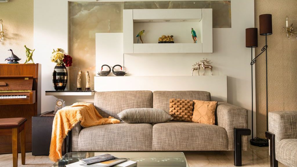 Warm living room with a gray sofa, mustard throw, textured pillows, floor lamps, wall shelving, and a glass coffee table.
