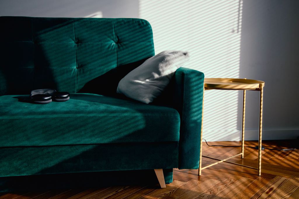 Deep green velvet sofa with tufted back cushions, a gold side table, and striped sunlight across a wood floor.