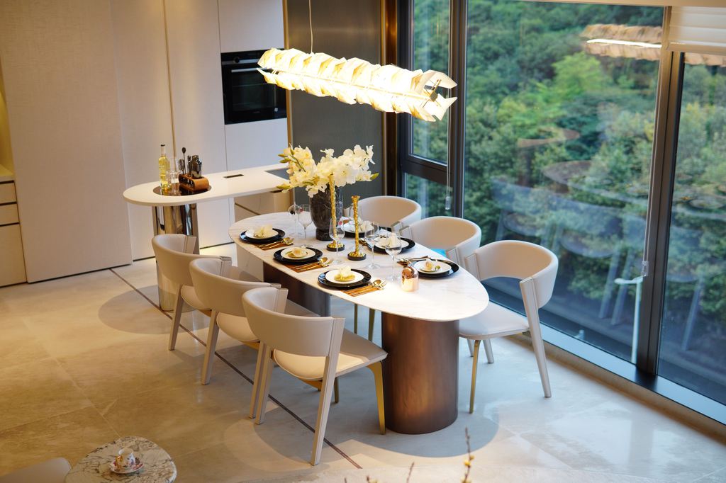 Oval designer dining table with a white top, curved pedestal base, six upholstered chairs, and a sculptural pendant light beside floor-to-ceiling windows.