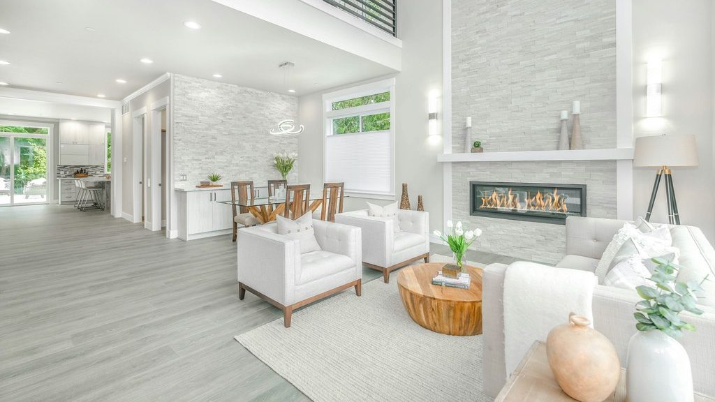 Bright white living room with a stone fireplace, cream seating, a natural wood coffee table, light gray flooring, and an open dining area.