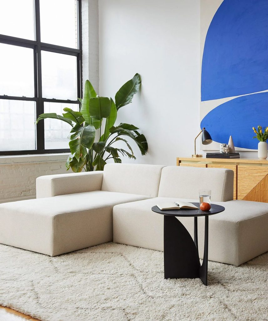 White low back modular sectional sofa in a bright modern living room with large windows, plants, and blue wall art.