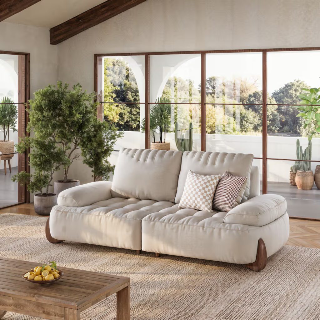 Beige low back sofa with tufted cushions and rounded arms in a bright living room with large windows and indoor plants.