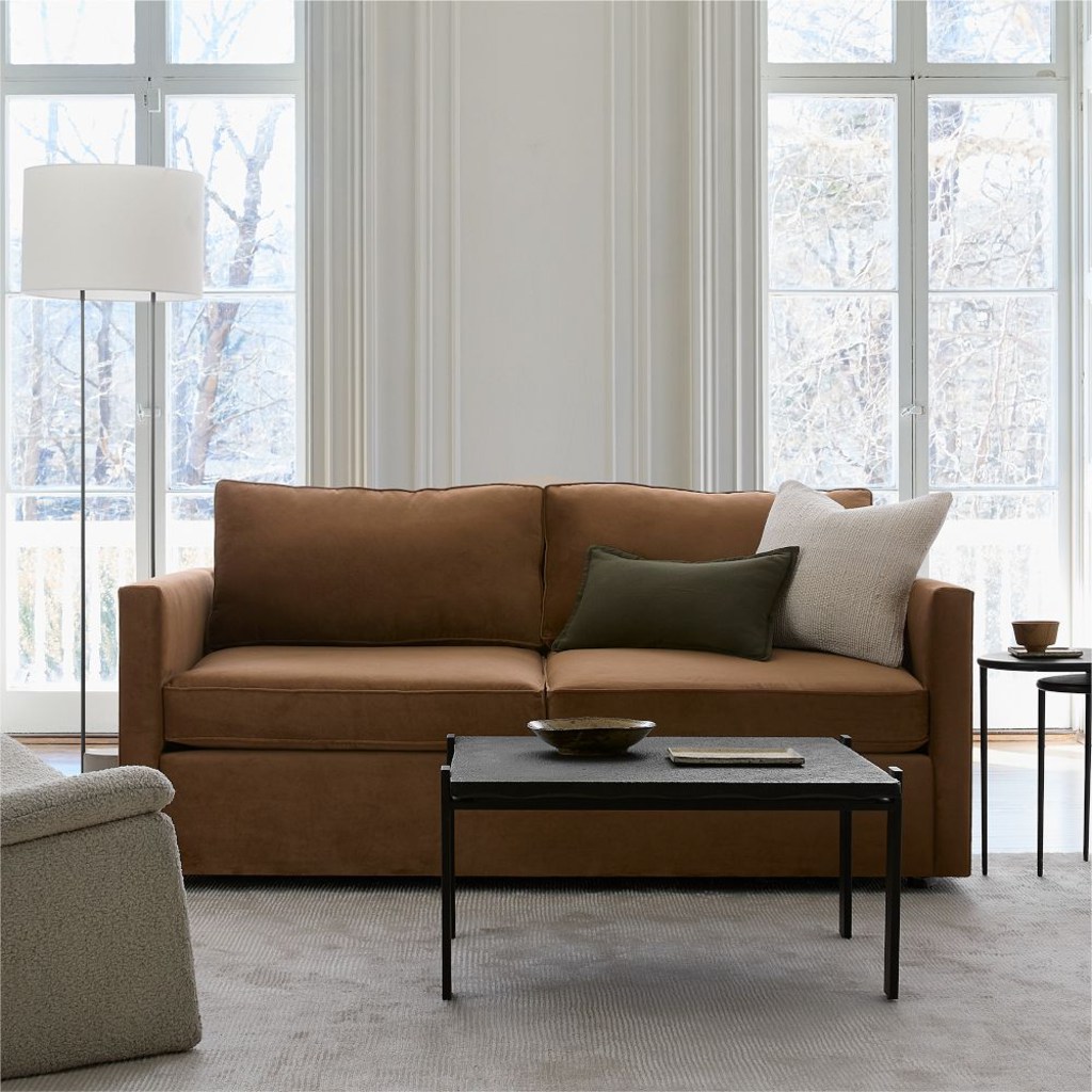 Brown upholstered sleeper sofa in a bright living room with large windows, neutral pillows, a black coffee table, and a floor lamp.