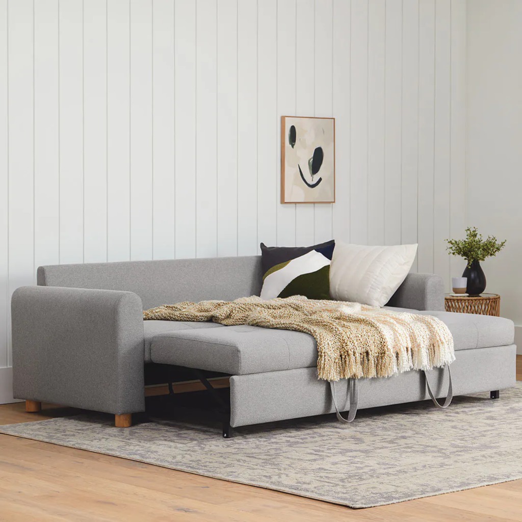 Gray slide-out sleeper sofa partially extended in a bright living room with pillows, a throw blanket, and a patterned rug.