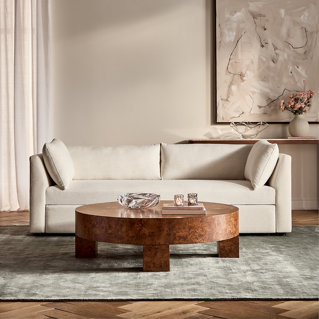 Cream upholstered sofa in an elegant neutral living room with a round wood coffee table, abstract wall art, and soft curtains.