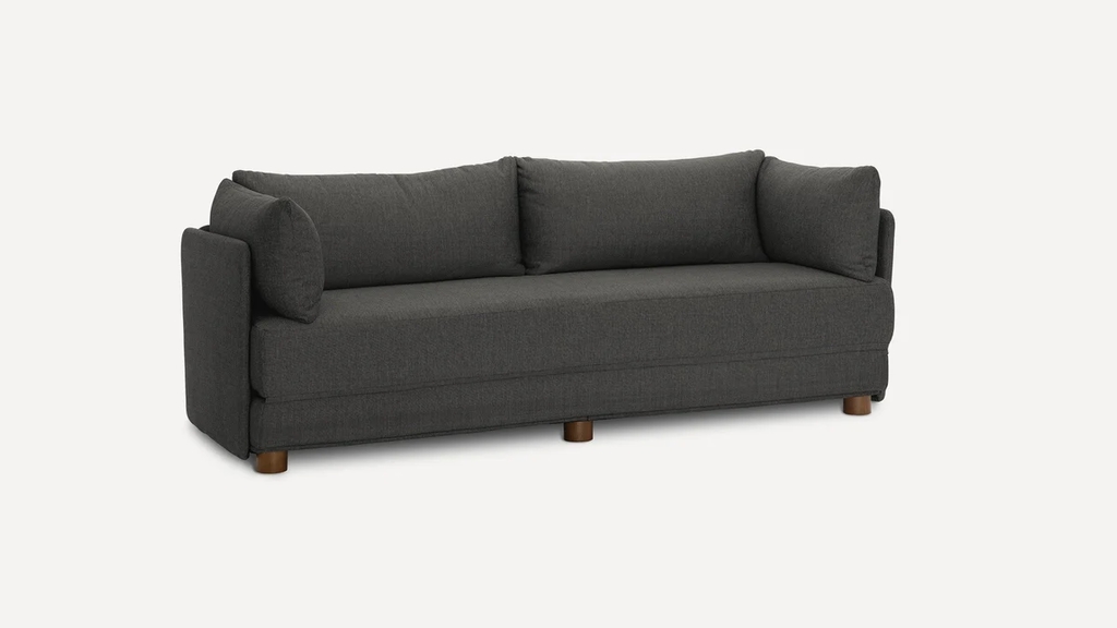 Dark gray sleeper sofa with plush back cushions, side pillows, and short wooden legs on a white background.