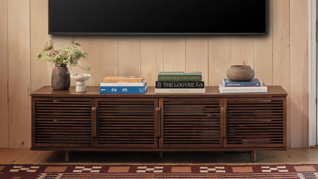 Wall-mounted TV above a dark walnut slatted media console with closed storage, books, flowers, and decorative objects.