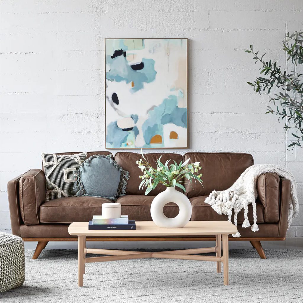 Brown leather sofa with raised wooden legs, decorative pillows, and a throw blanket in a bright modern living room.