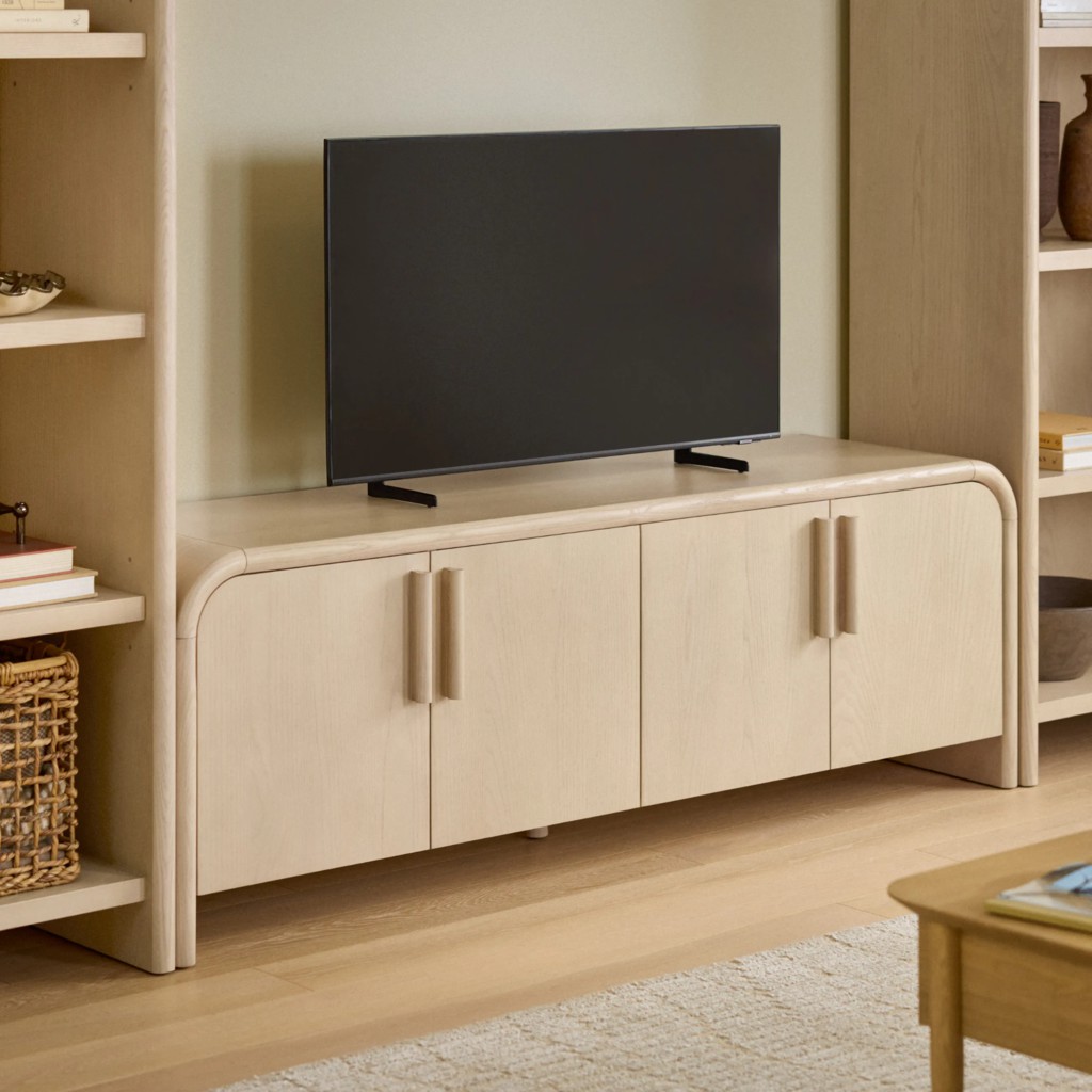 Light wood media console with rounded corners and cabinet doors, supporting a flat-screen TV between built-in shelving in a warm neutral living room.