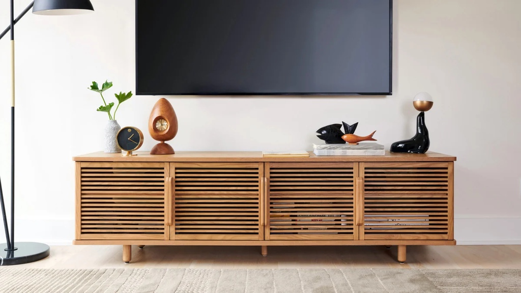 Light wood slatted media console beneath a wall-mounted TV, styled with clocks, books, decorative objects, and a floor lamp in a bright modern living room.
