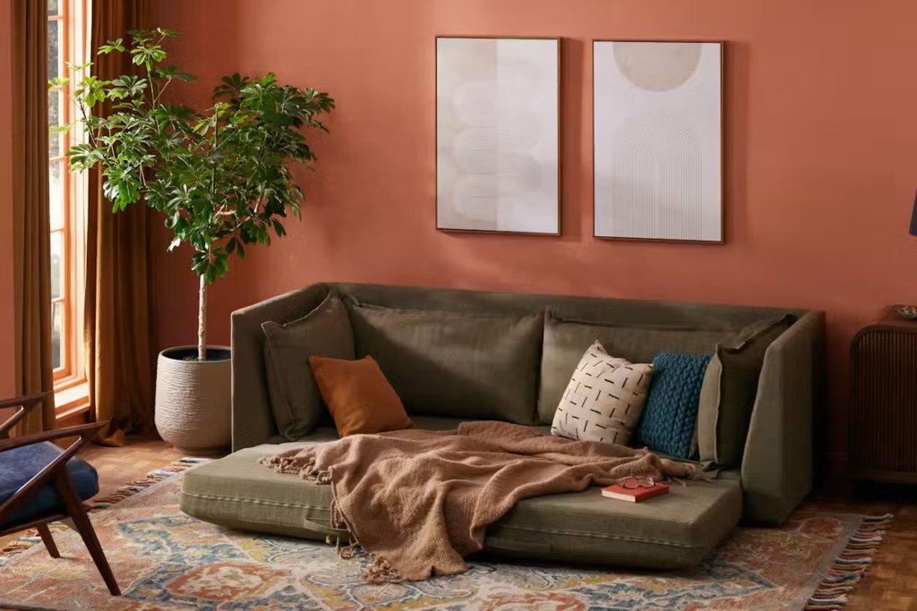 Olive green fold-out sofa bed opened into a sleeping platform in a warm living room with terracotta walls, plants, pillows, and a patterned rug.