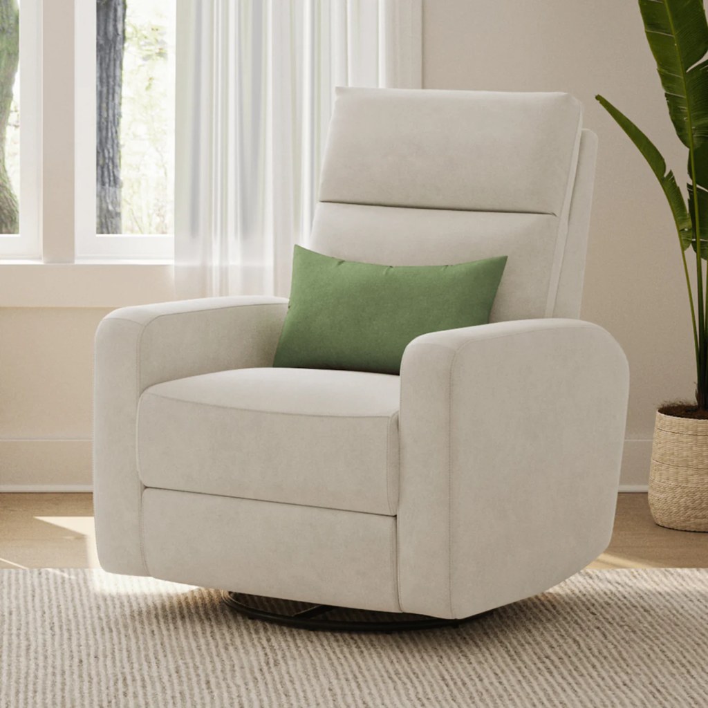 Cream upholstered swivel nursery recliner with a green lumbar pillow beside a bright window and potted plant.