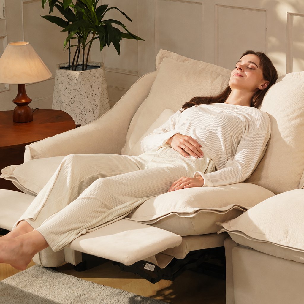 Woman relaxing on a cream reclining sofa with extended footrest in a warm living room with a side table, lamp, and indoor plant.