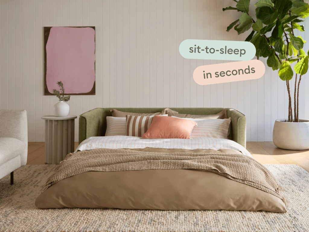 Green sofa bed opened into a sleeping platform with neutral bedding in a bright modern room with plants and wall art.