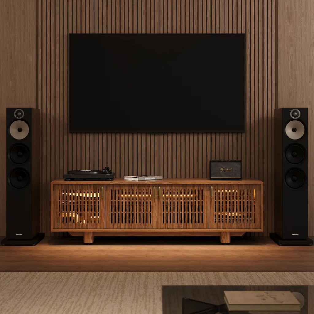 Wood slatted media console beneath a wall-mounted TV, flanked by floor speakers in a home audio setup.