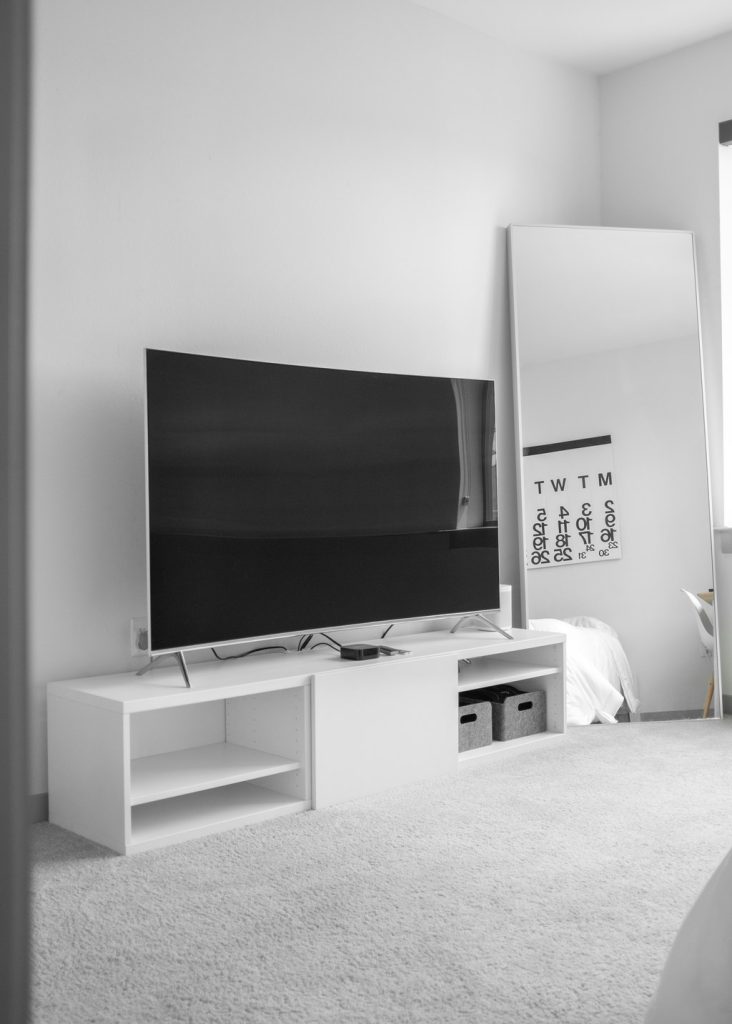 Flat-screen TV on a white media console with open shelving in a minimalist bedroom.
