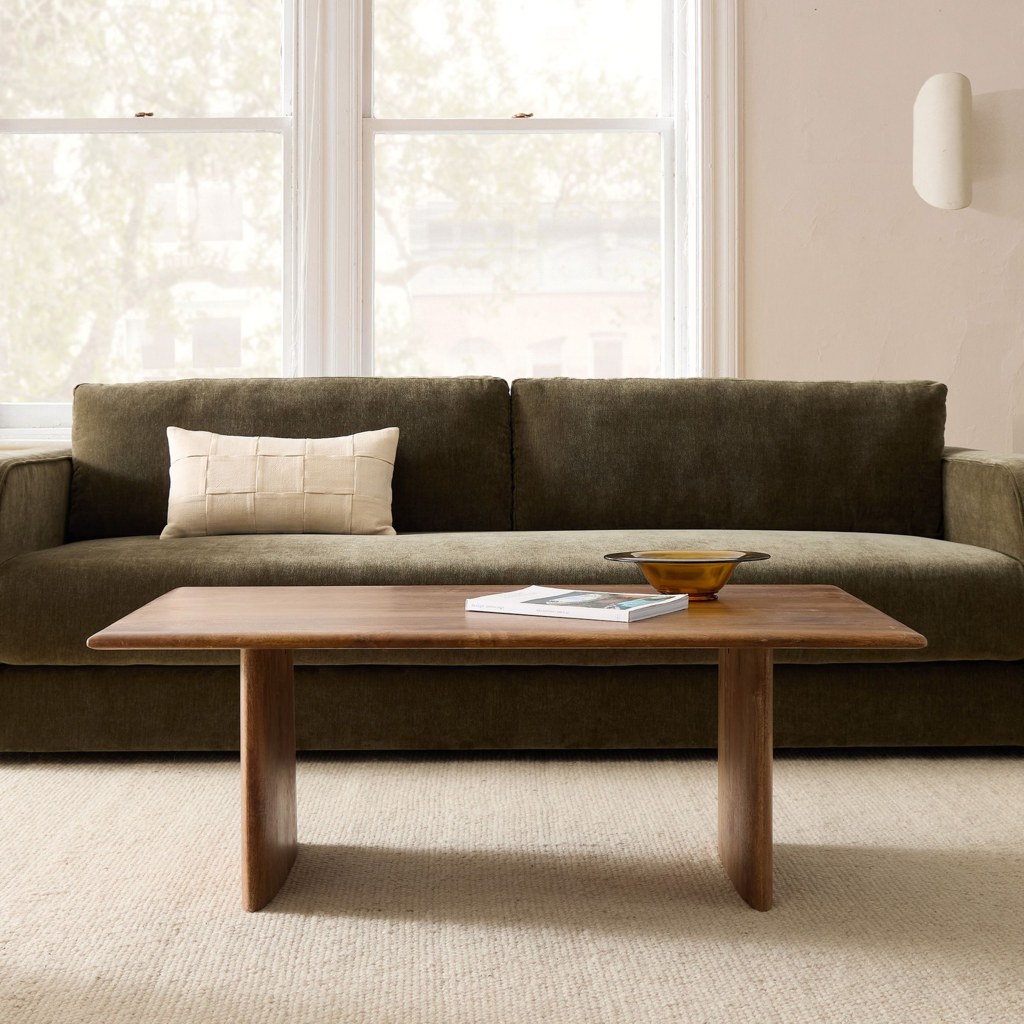 Wooden coffee table in front of a dark green sofa in a bright minimalist living room with a book and decorative bowl on the tabletop.