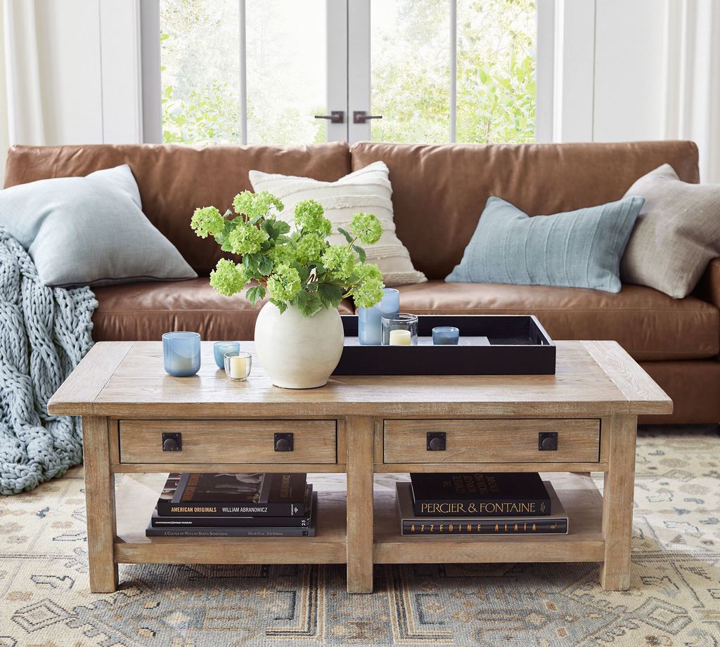 Rectangular wood coffee table with drawers and lower shelves in a cozy living room featuring a leather sofa, books, and decorative accents.