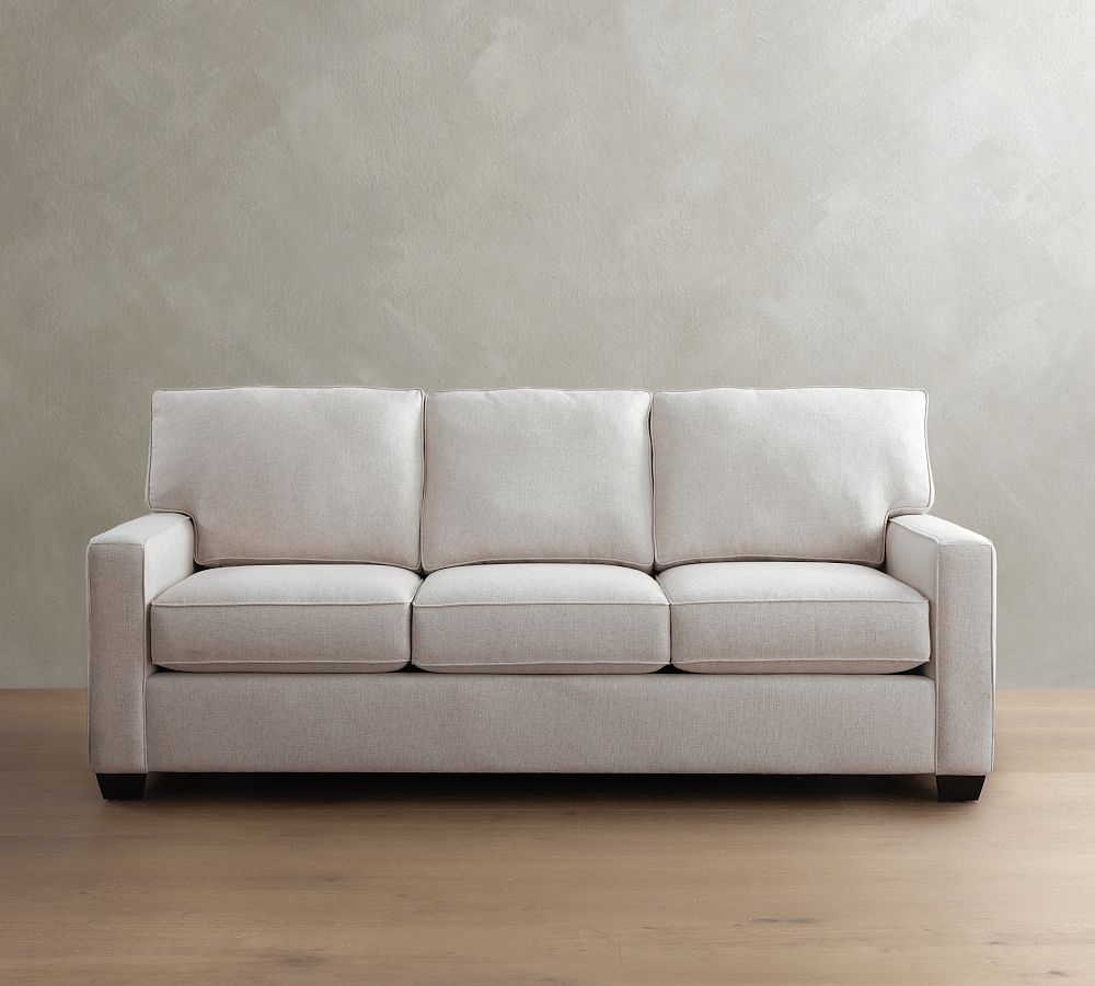 Light beige sleeper sofa with three seat and back cushions against a neutral wall on a wood floor.
