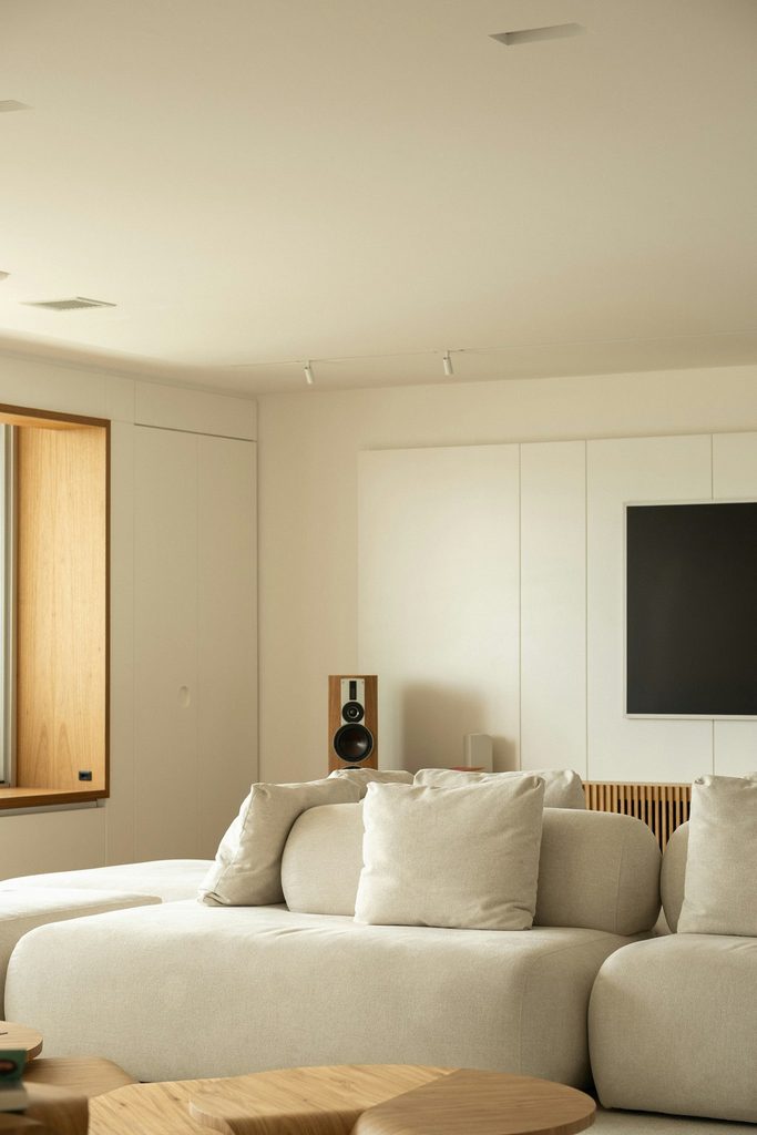 Modern living room with a cream sectional sofa, round wooden coffee table, wall-mounted TV, and minimalist neutral interior design.