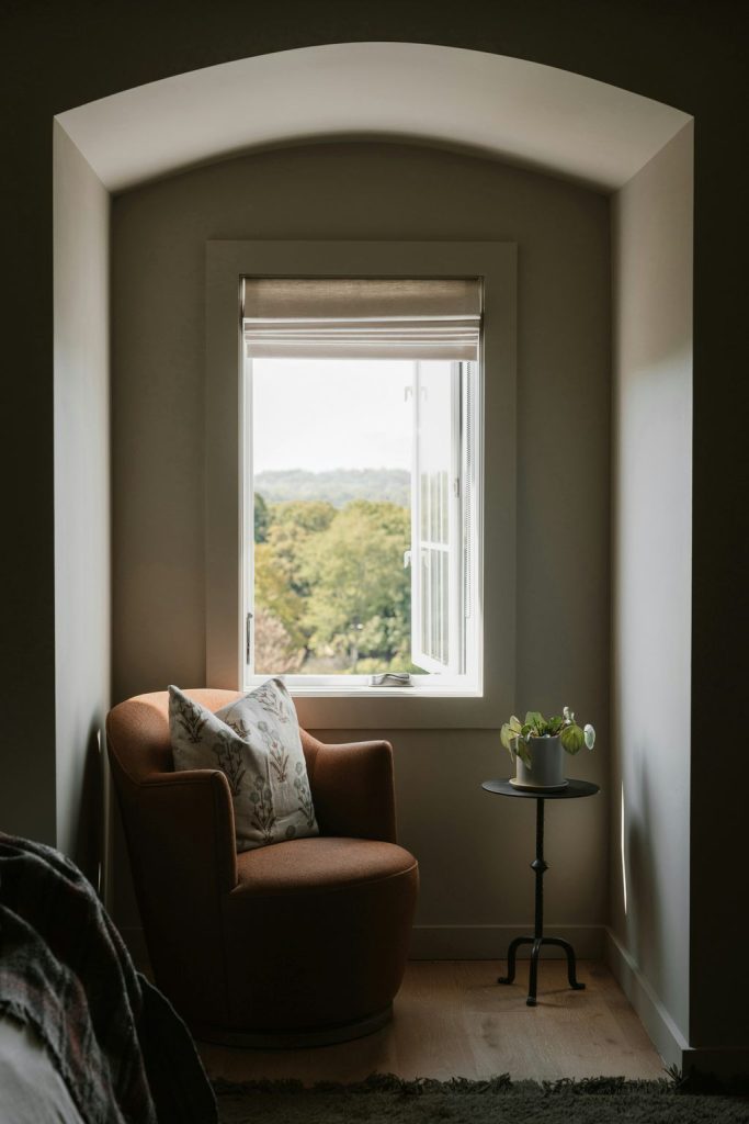 Rust-colored armchair with a floral pillow beside a small table in a cozy window nook.