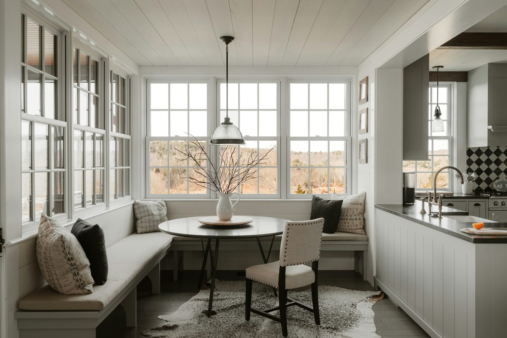 Bright breakfast nook with built-in bench seating, round dining table, and large windows beside a modern kitchen.