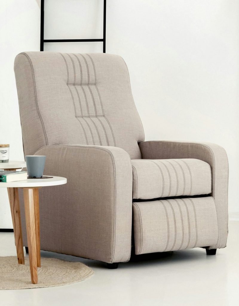 Beige upholstered recliner chair beside a small round table in a bright minimalist room.