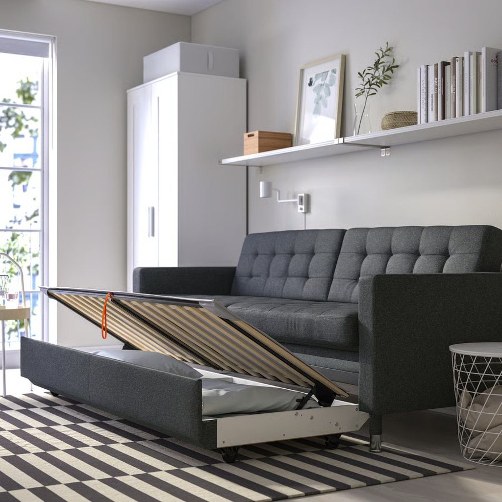Dark gray sleeper sofa with a pull-out slatted bed frame extended in a compact modern room with shelving, wardrobe, and striped rug.