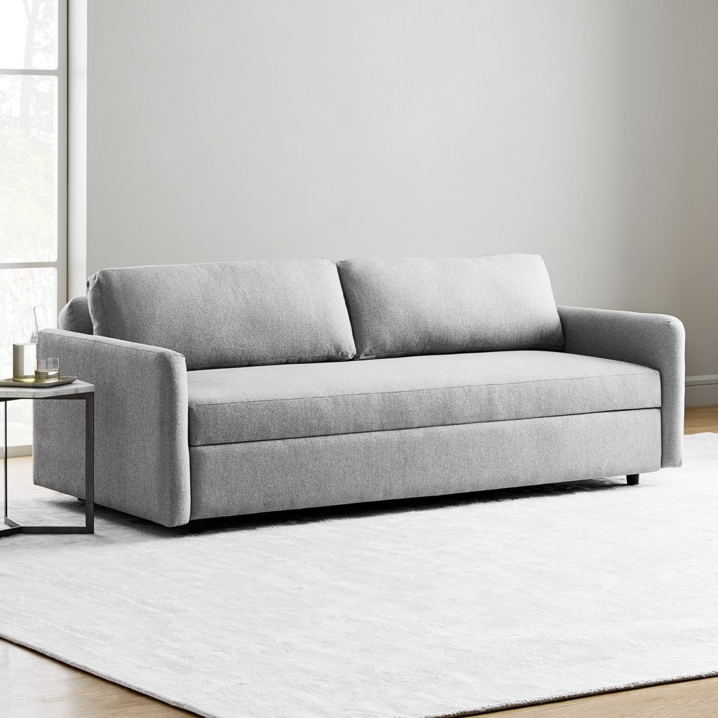Light gray sleeper sofa with a bench seat and two back cushions in a bright minimalist living room with a white rug and side table.