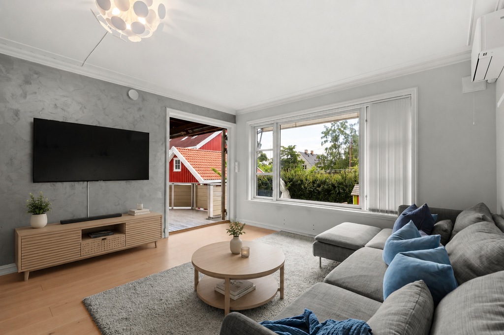 Bright modern living room with a wall-mounted TV, light wood TV stand, round coffee table, gray sectional sofa, and large window.