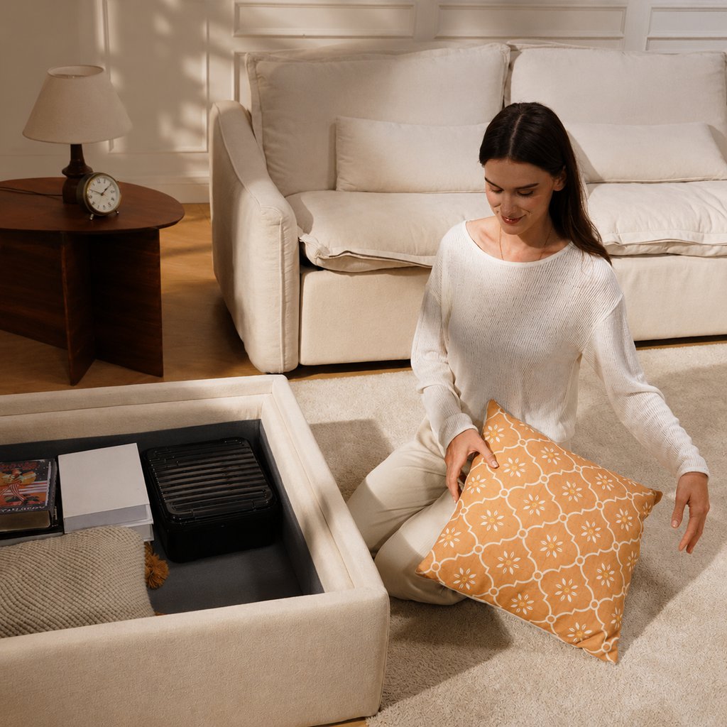 Woman organizing pillows beside an open storage ottoman in a warm neutral living room.