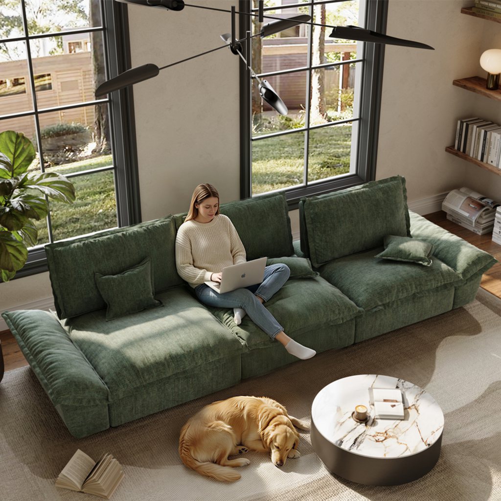 Green deep-seat sofa in a sunlit living room with a woman using a laptop and a dog resting nearby.