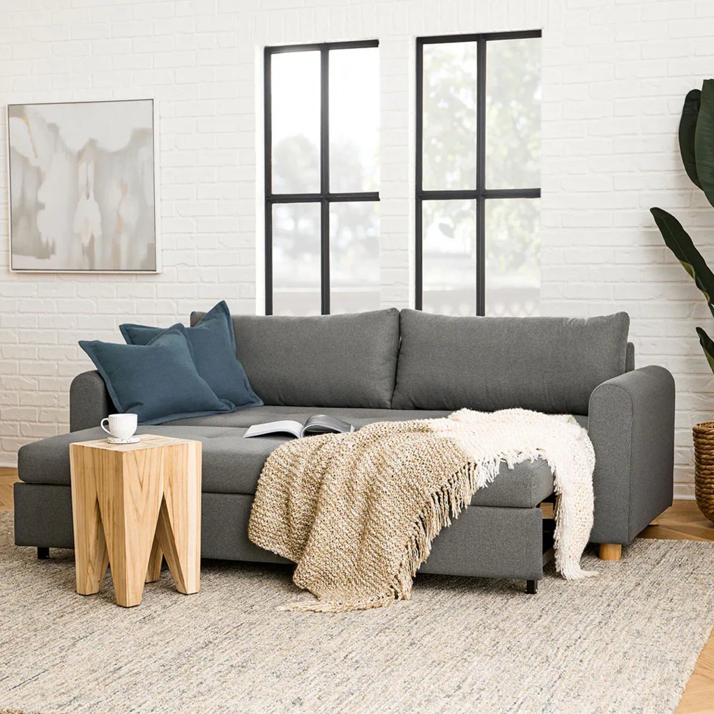 Gray sleeper sofa with a pull-out chaise, layered throws, blue accent pillows, and a side table in a bright modern living room.
