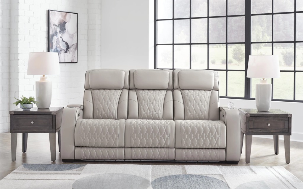 Light gray three-seat power reclining sofa with quilted upholstery in a bright living room with large windows and side tables.