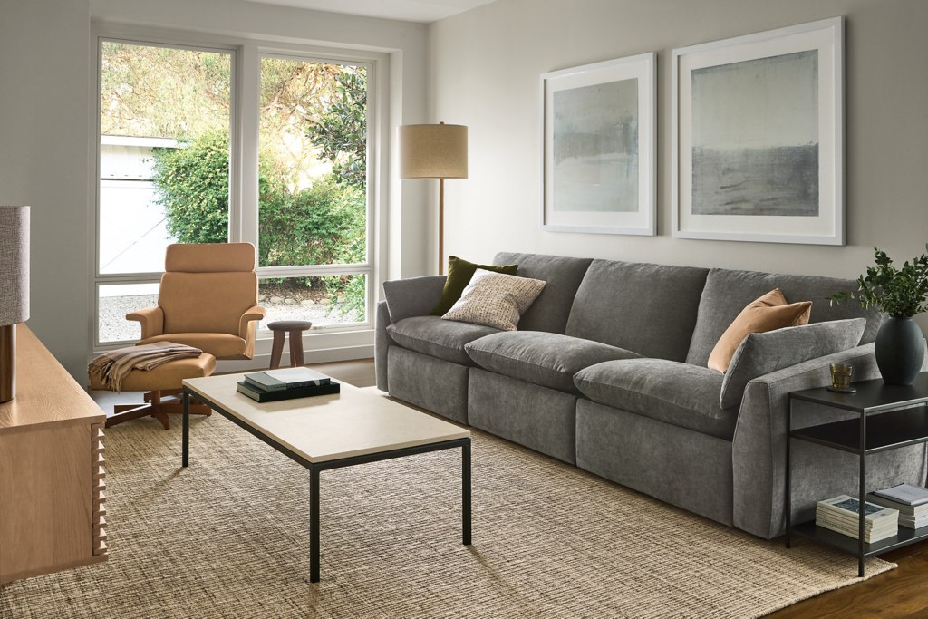 Gray three-seat power reclining sofa in a modern living room with a lounge chair, coffee table, and large windows.